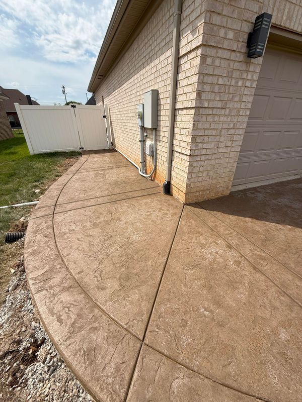 Concrete pathway curves along a brick building, leading to a garage door. Brown surface, white fence, and greenery visible.