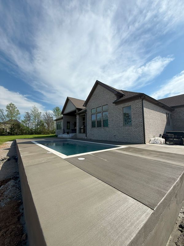 Concrete patio with a rectangular pool and a house in the background under a blue sky.