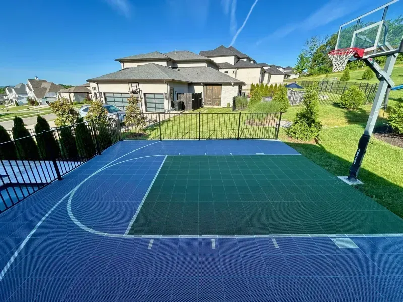 Blue and green basketball court with hoop, set in a backyard near a house and green lawn under a sunny, blue sky.