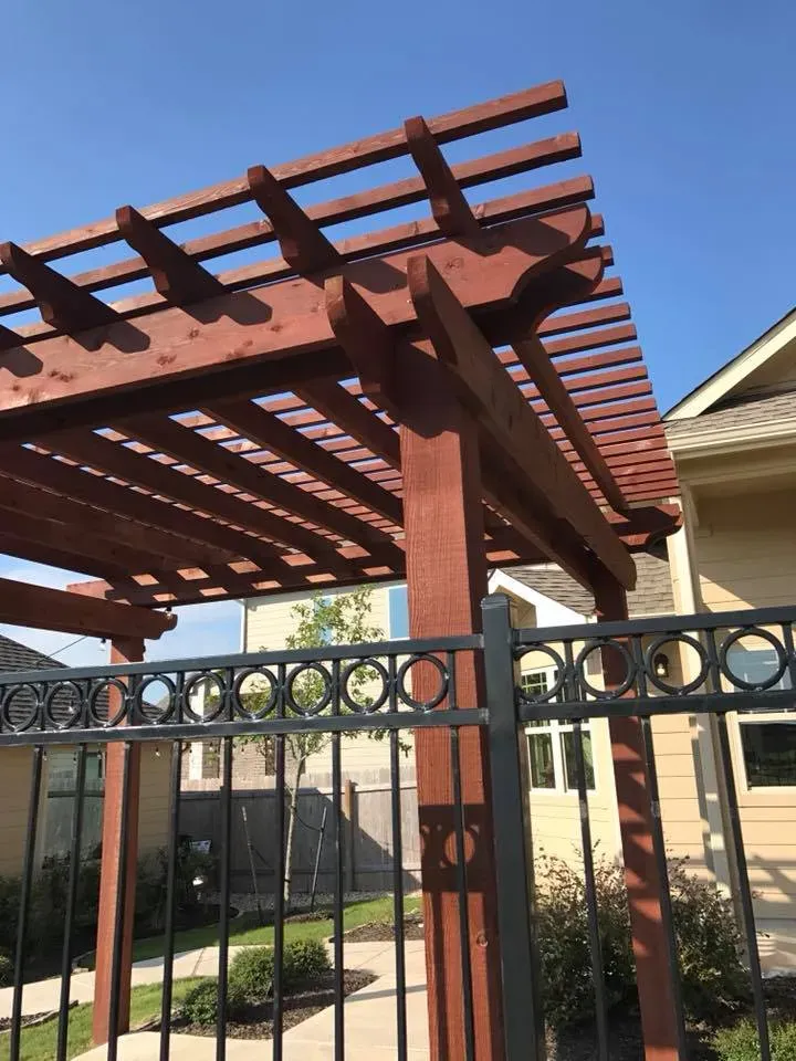 A wooden pergola is behind a metal fence in front of a house.