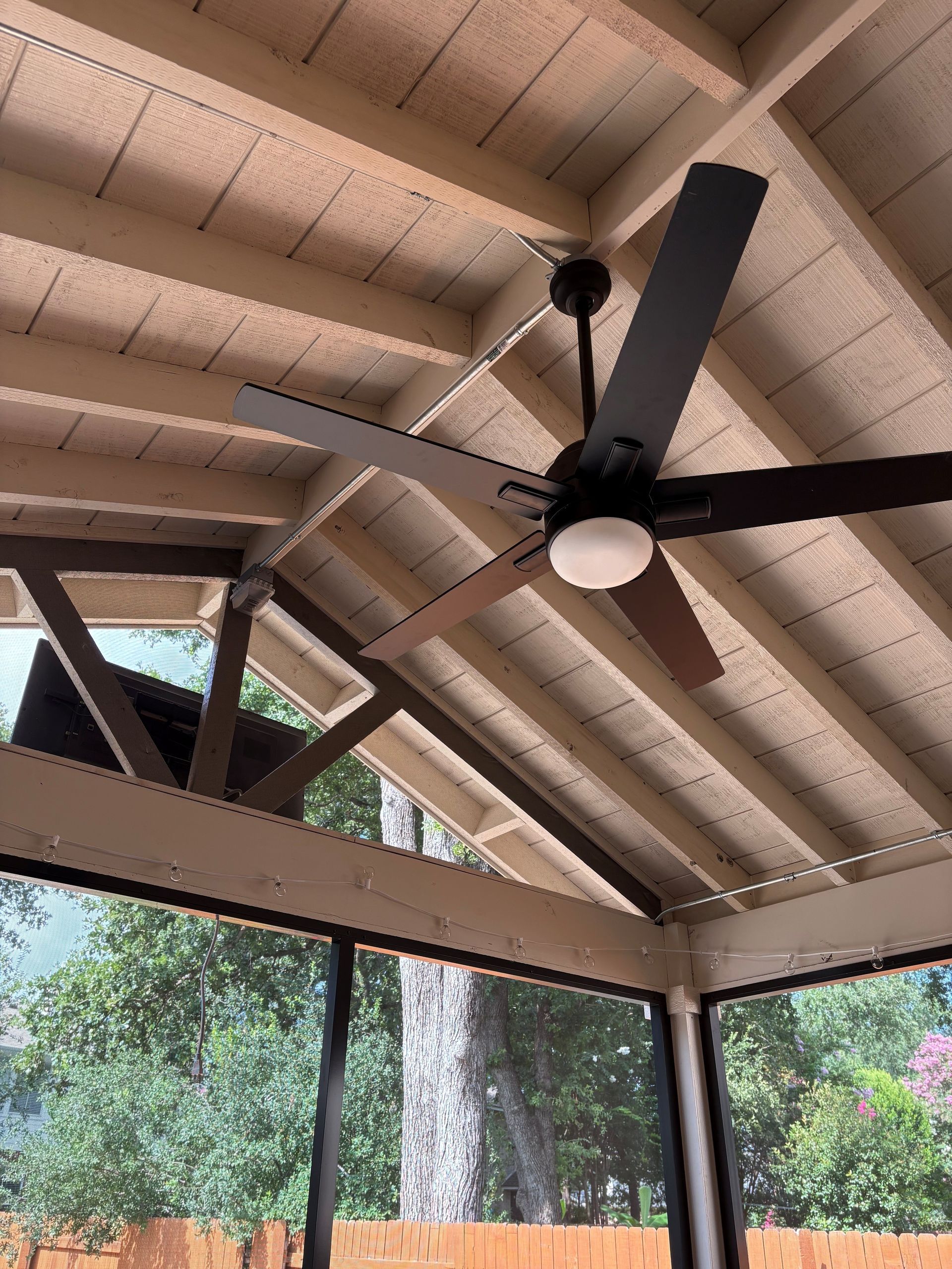 Covered patio ceiling with a black ceiling fan and tan beams, overlooking trees outside