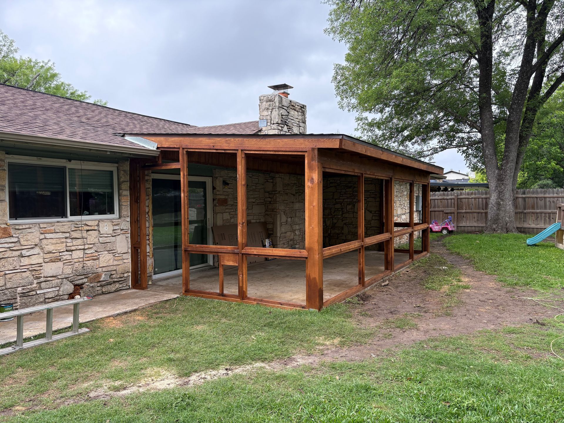 Backyard patio with a wooden screened porch extension beside a stone house and grassy yard.