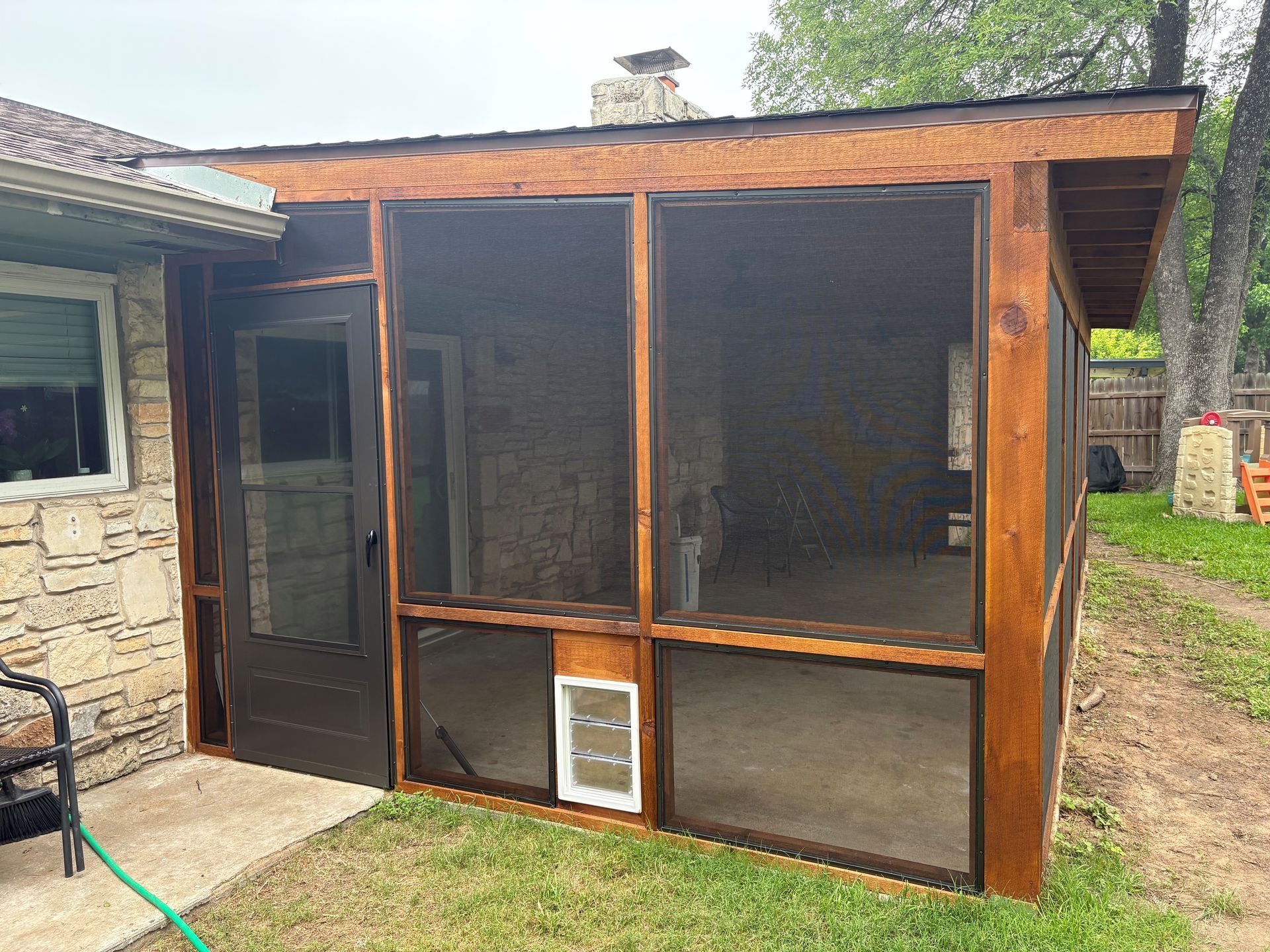 Wooden outdoor screened enclosure attached to a stone house, with mesh panels and a small pet door.