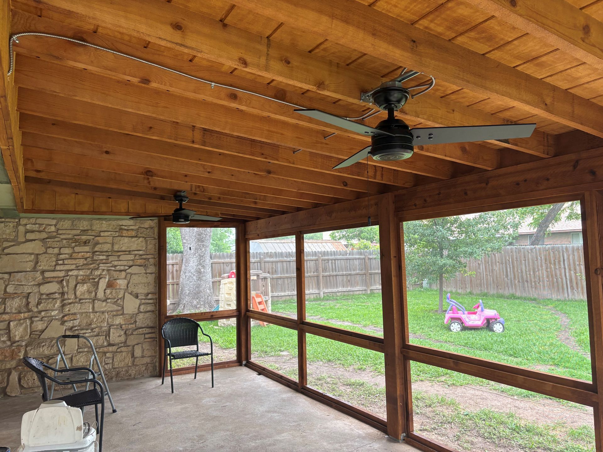 Covered patio with wood ceiling, stone wall, ceiling fan, and yard view with a pink toy car