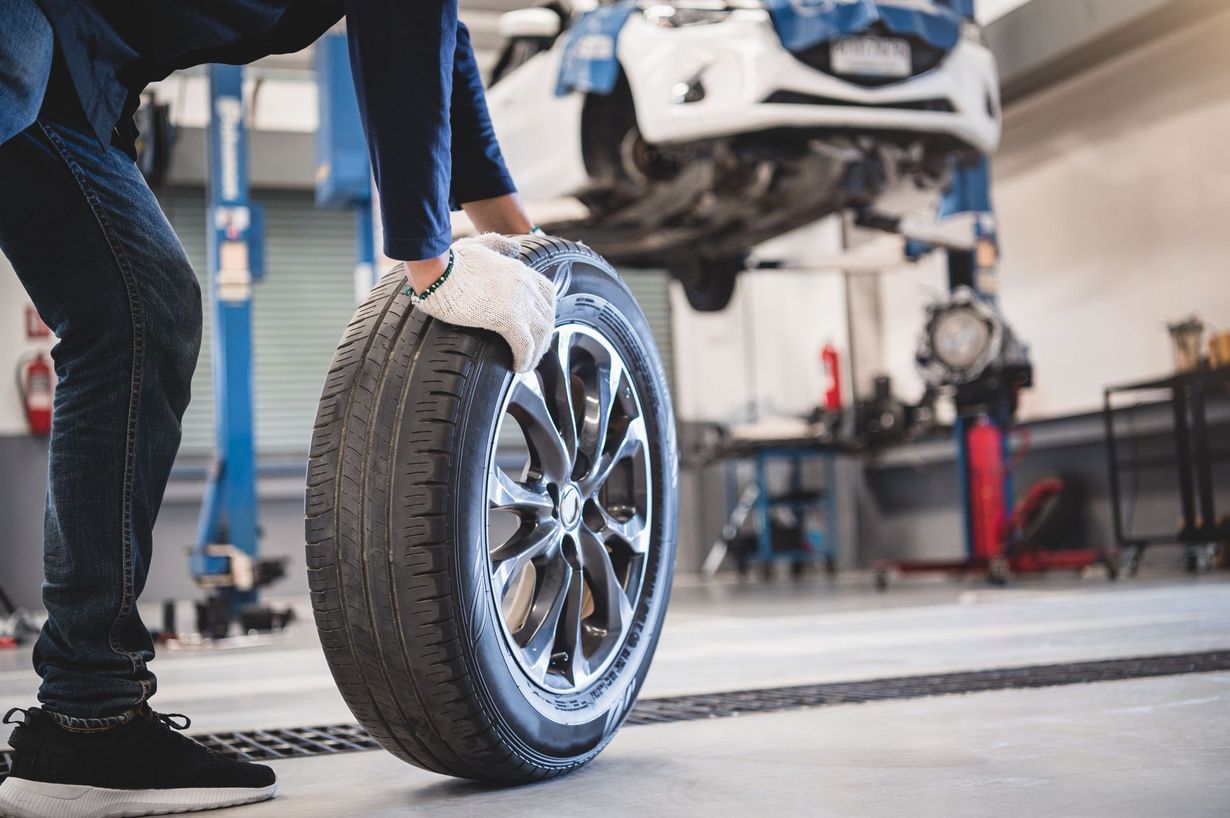 Mechanic rolling a car tire in a repair shop with a car lifted in the background