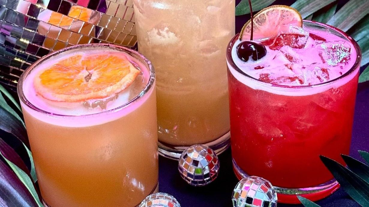 Three cocktails with garnishes, including one red drink with a cherry, surrounded by small disco balls on a dark surface.