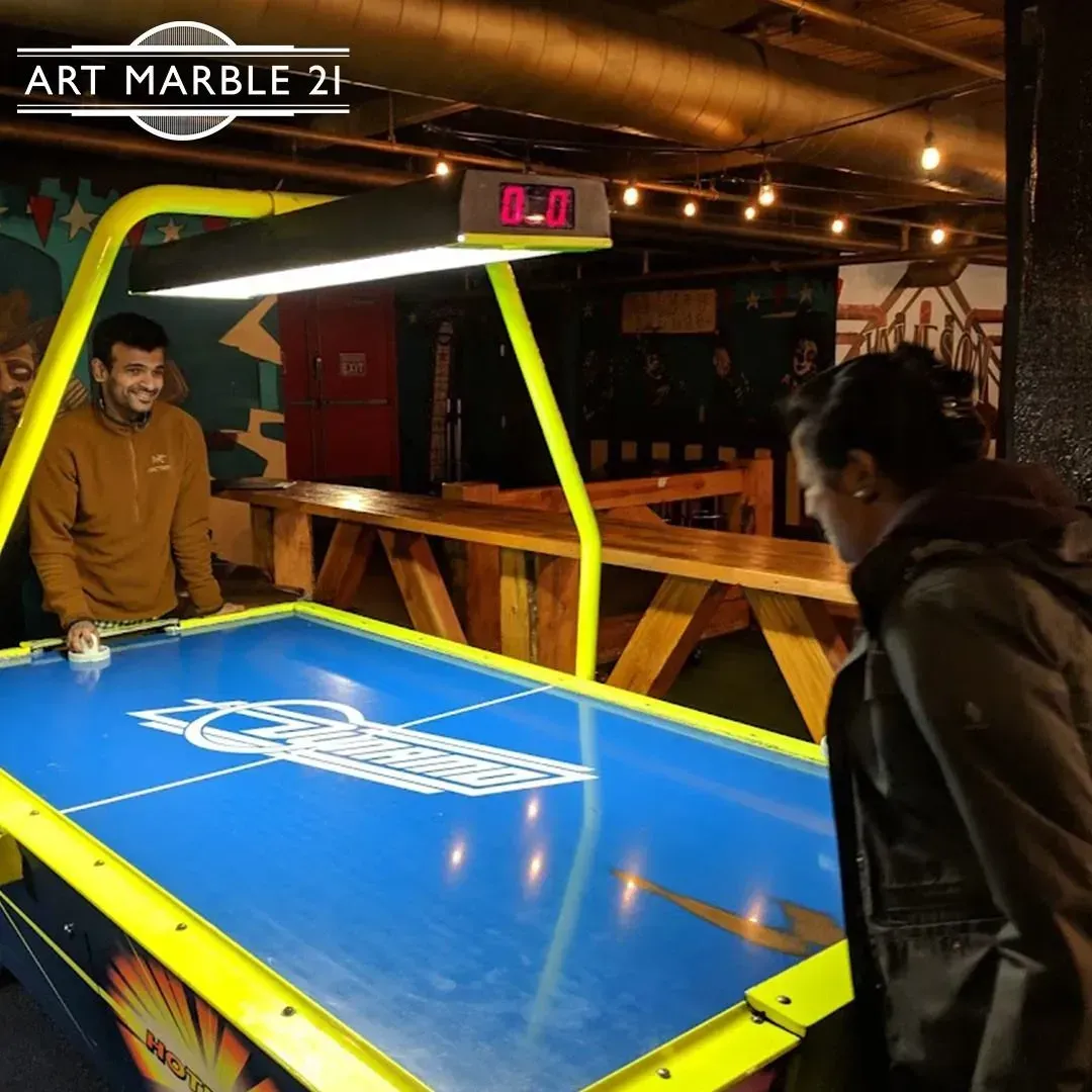 Two people playing air hockey at a bar; blue table, yellow frame, digital score display, warm lighting.