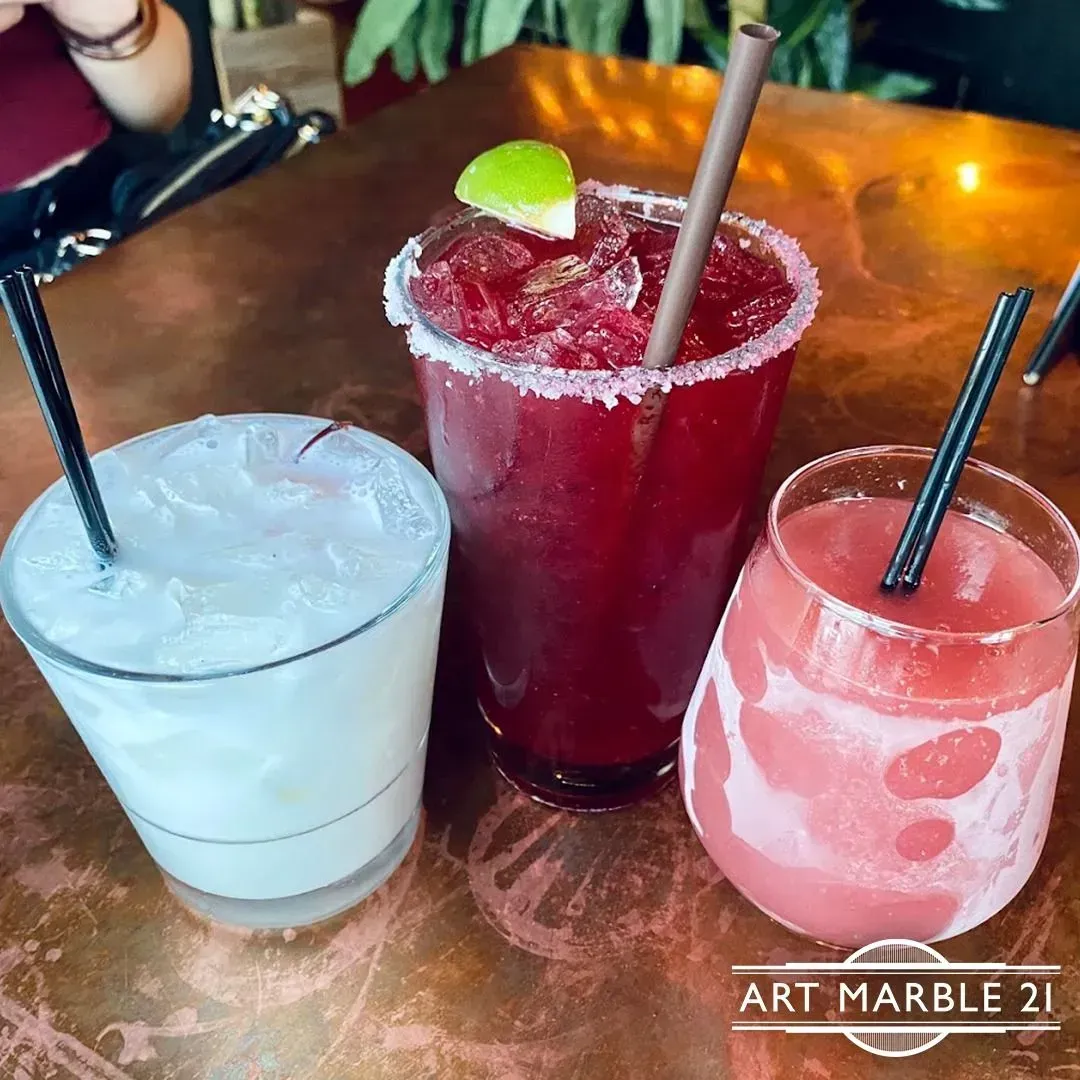 Three colorful cocktails on a copper table, one white, one red, and one pink, each with a straw.