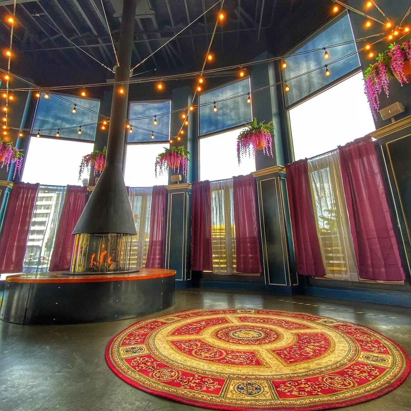 Cozy, circular room with a fireplace, large windows, and a red rug, adorned with string lights.