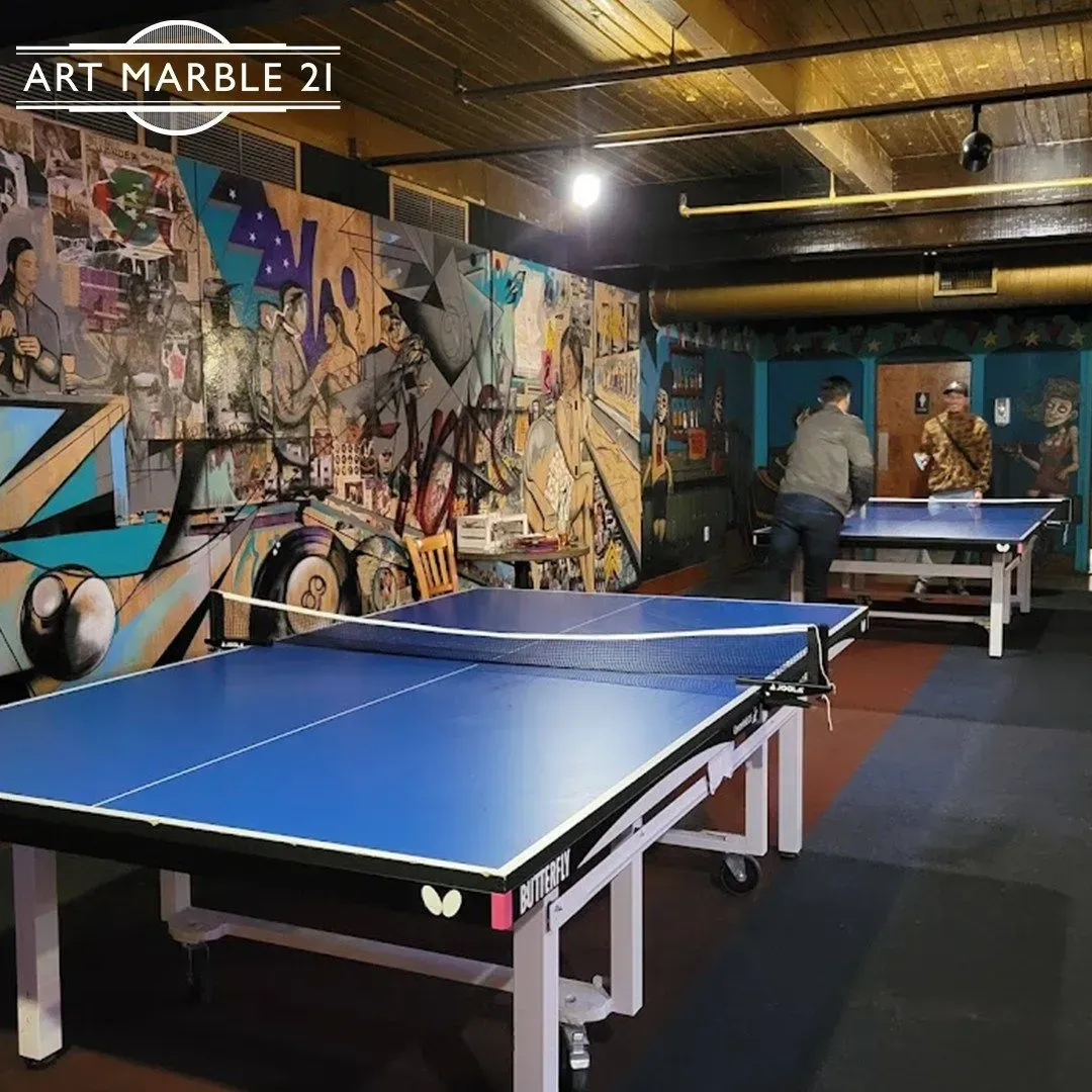 Two ping pong tables in a room with a mural on the wall, two people playing.