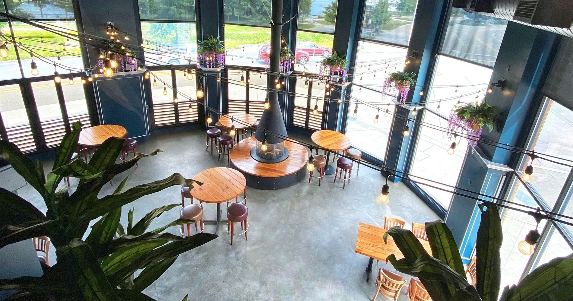 High-angle view of a modern lounge with a central fireplace, wooden tables, and hanging potted plants by large windows.