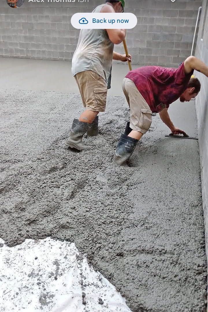 Two workers in boots smoothing fresh wet concrete with a long tool indoors.