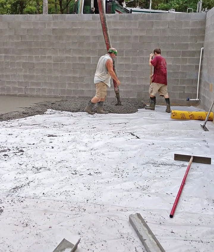 Workers leveling wet concrete in a backyard beside a cinder block wall