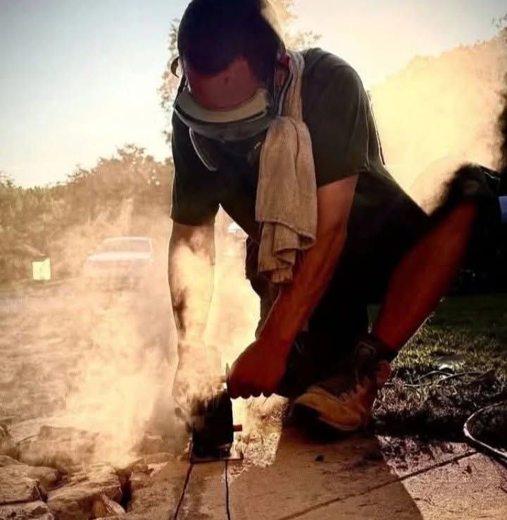 Worker using a power tool cuts concrete outdoors, with dust and debris around the job site.