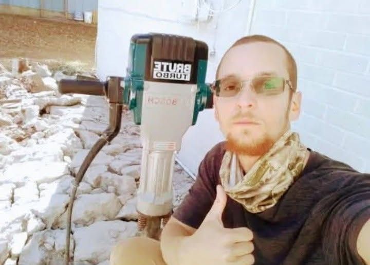 Man taking a selfie beside a large survey instrument in a rocky outdoor work site