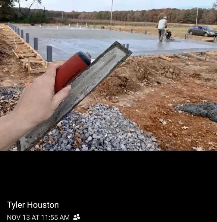 Hand holding a red and gray trowel beside a freshly poured concrete slab outdoors