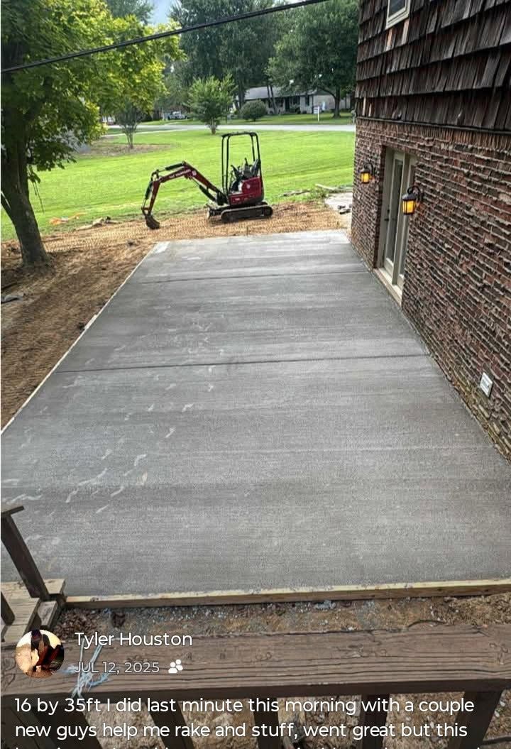 Fresh concrete driveway beside a brick house, with a small excavator at the far end and a grassy yard beyond.