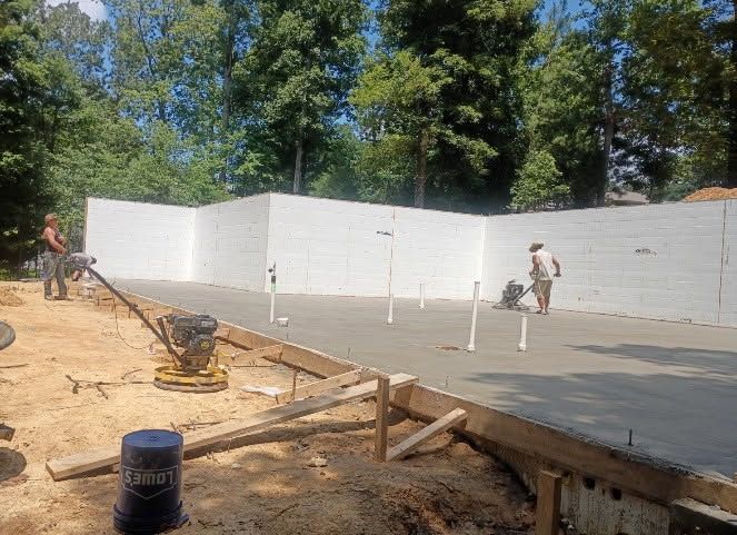 Construction site with workers pouring a concrete slab beside white foundation walls and trees.