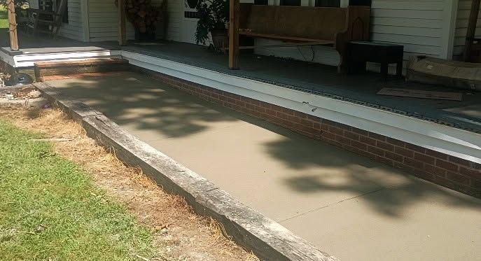 Concrete ramp leading to a porch with white railing and brick foundation in a shaded yard