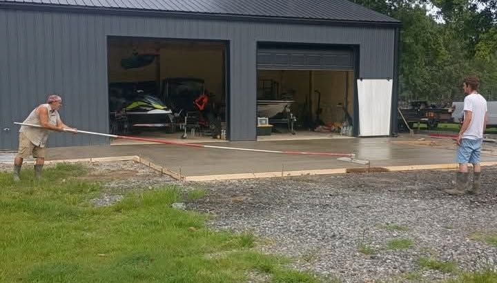 Two people stretch a tape across a gravel driveway in front of a gray garage with open doors.