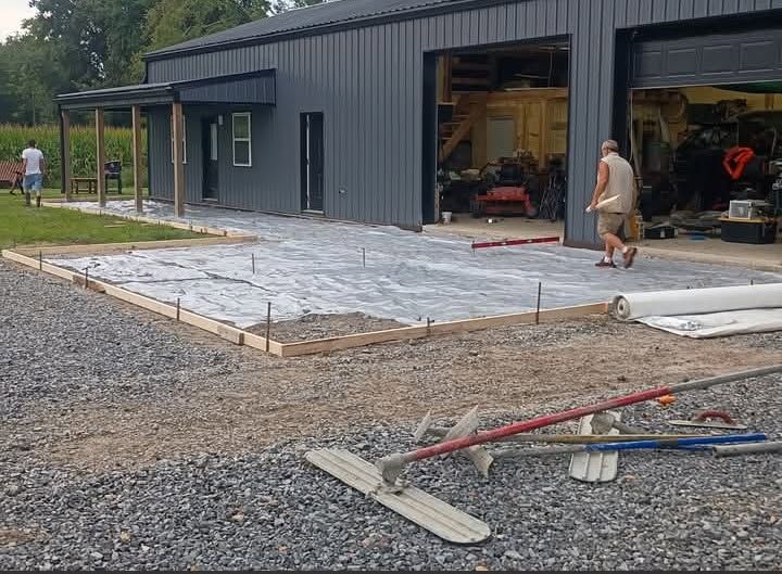 Unfinished concrete slab beside a dark metal workshop, with tools in the gravel and a person near the open garage.