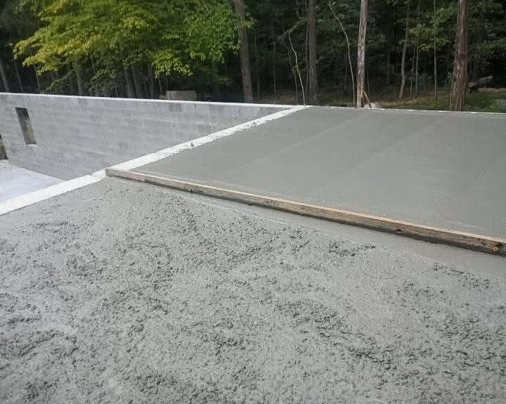 Freshly poured concrete slab beside a gravel surface and wooded area, with a wooden edge form in place