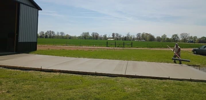 Rural scene with a concrete path beside a barn and a person walking a dog across a grassy field
