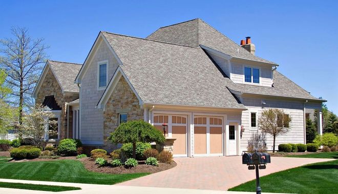 Two-story house with gray roof, tan brick and light-colored siding, two-car garage, and manicured lawn under a blue sky.