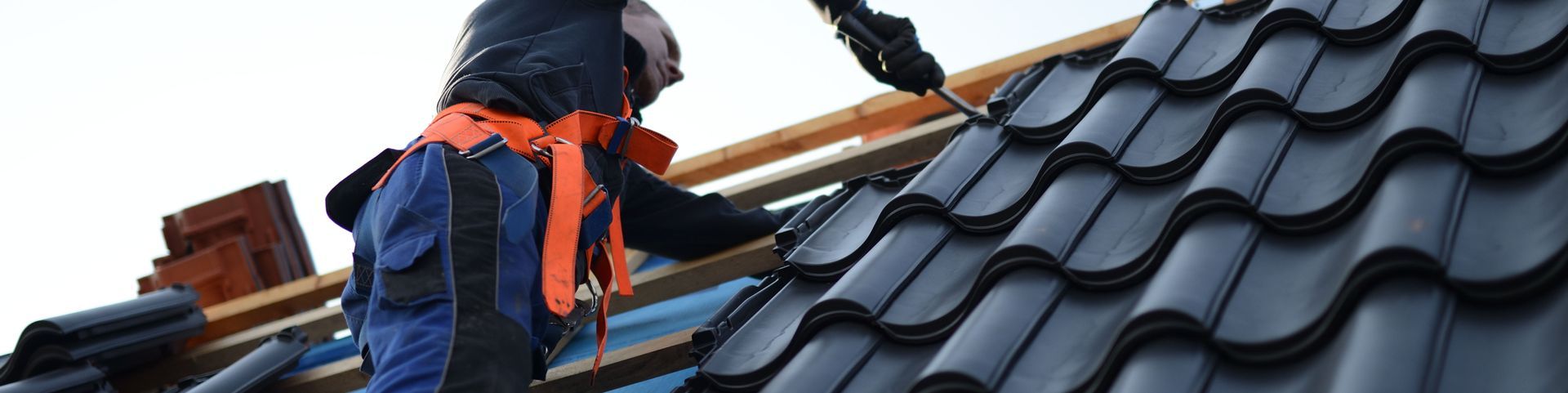 Roofer wearing safety harness working on a dark tiled roof, using a tool.