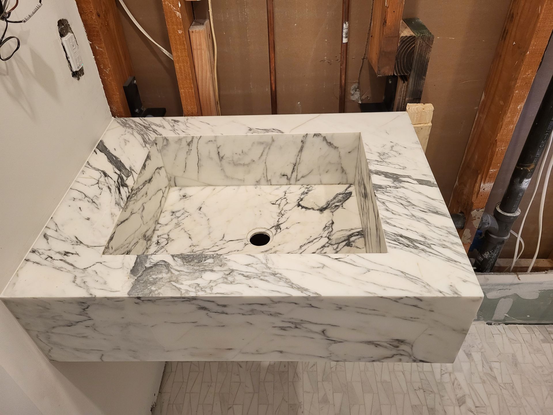 Marble, wall-mounted bathroom sink with square basin. White with grey veining, installed against a partially constructed wall.