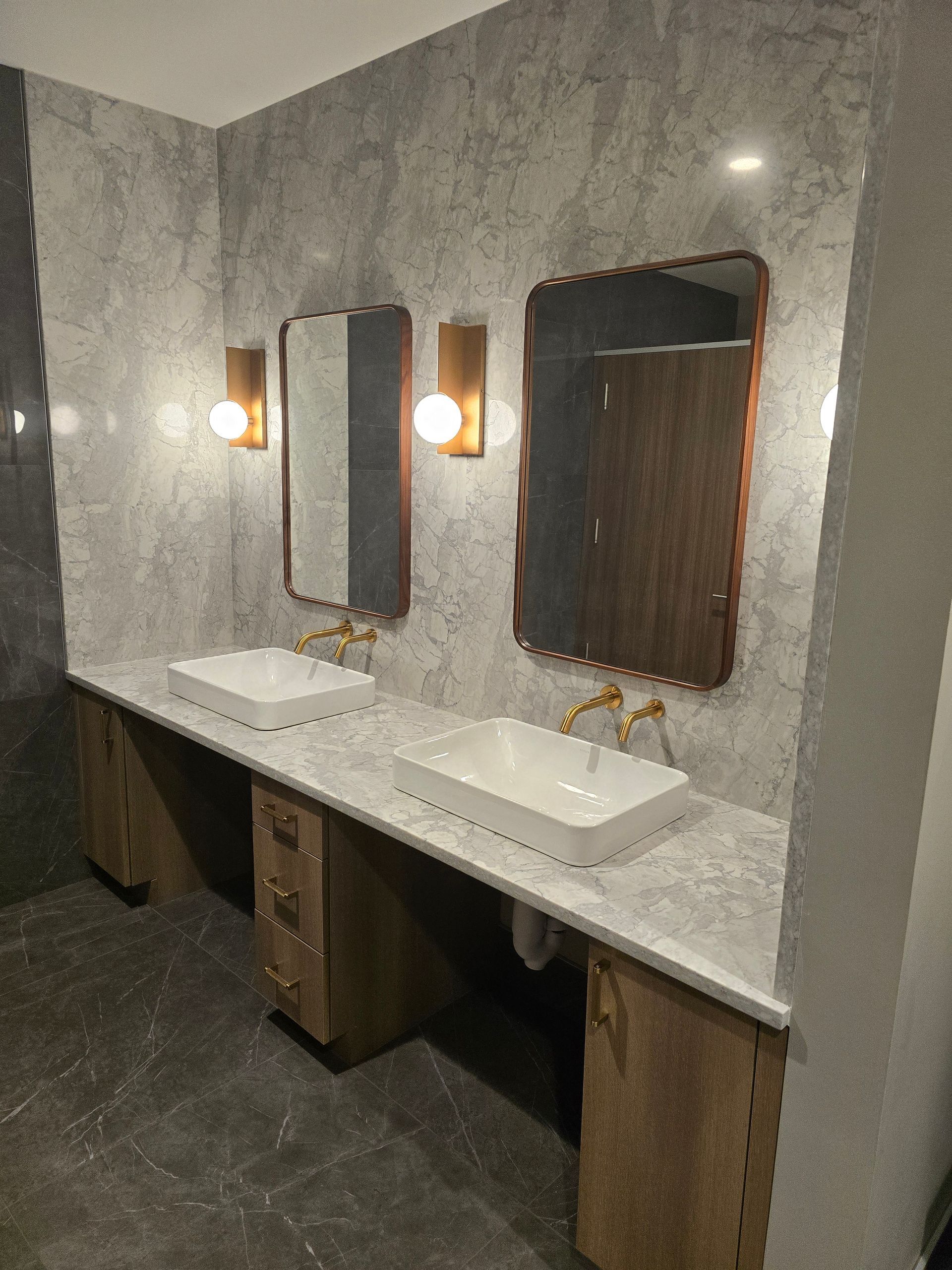 Bathroom with two sinks, two mirrors, gold fixtures, and wood cabinets against a gray stone wall and dark tiled floor.