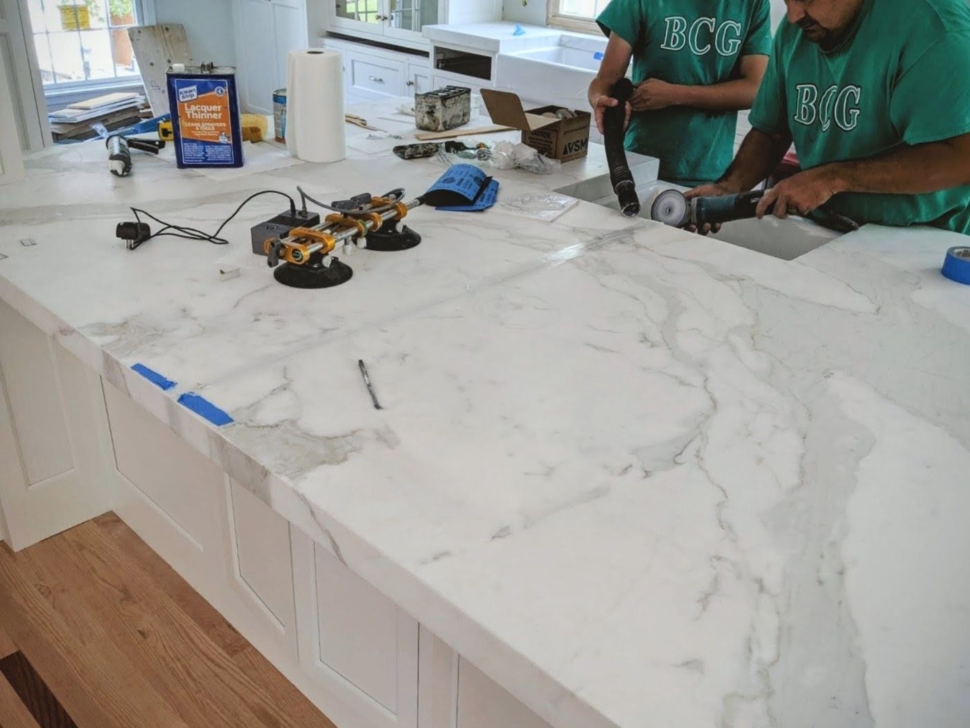 Two workers cutting a white marble countertop in a kitchen; white cabinets and wood floor visible.