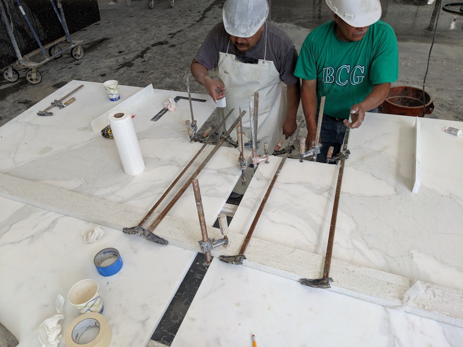Two workers in a workshop use tools on a large white marble slab, applying adhesive to a seam.
