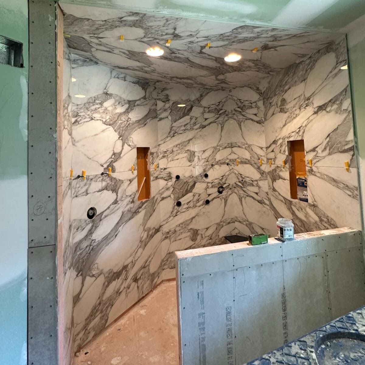 Shower stall under construction, featuring large marble-patterned tiles, with recessed shelves and overhead lights.