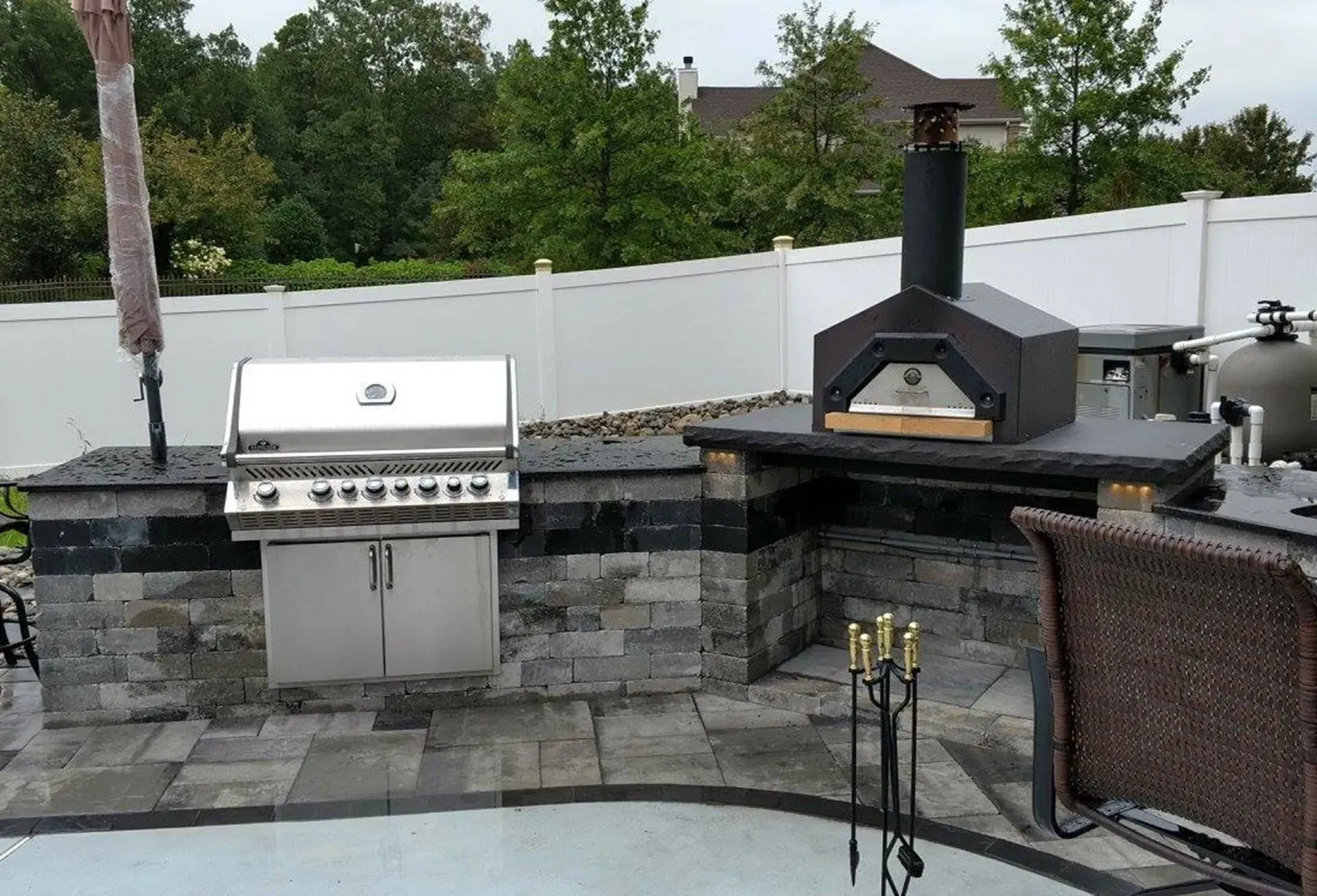 Outdoor kitchen with grill, pizza oven, and a white fence background.