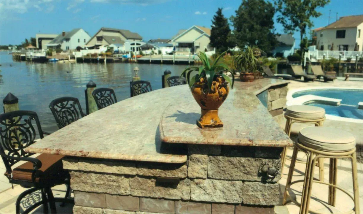 Outdoor bar with water view; granite countertop, stone base, chairs, pool, boats, houses in background.