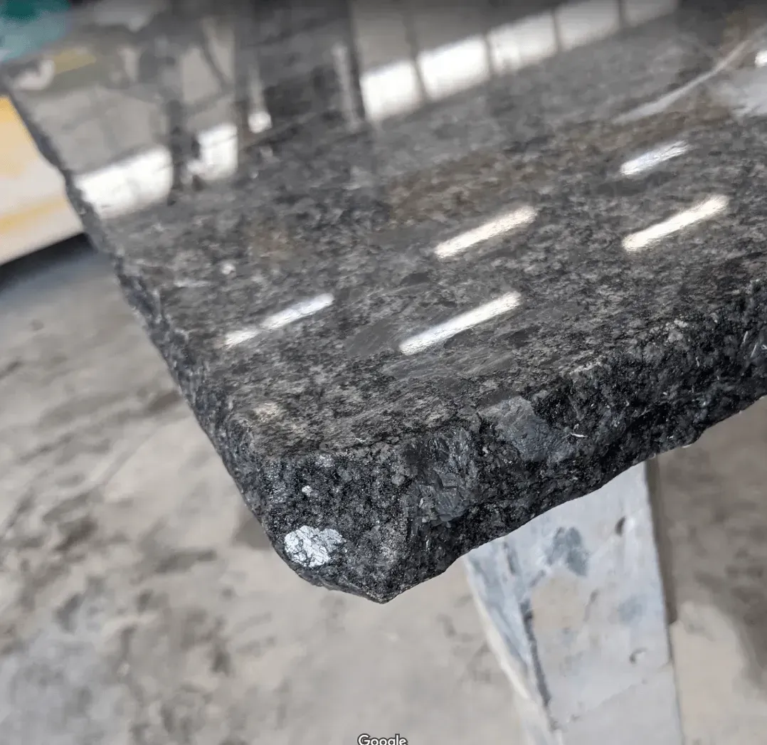 Close-up of a dark granite countertop with a rough, natural edge; shiny surface reflects bright lights.