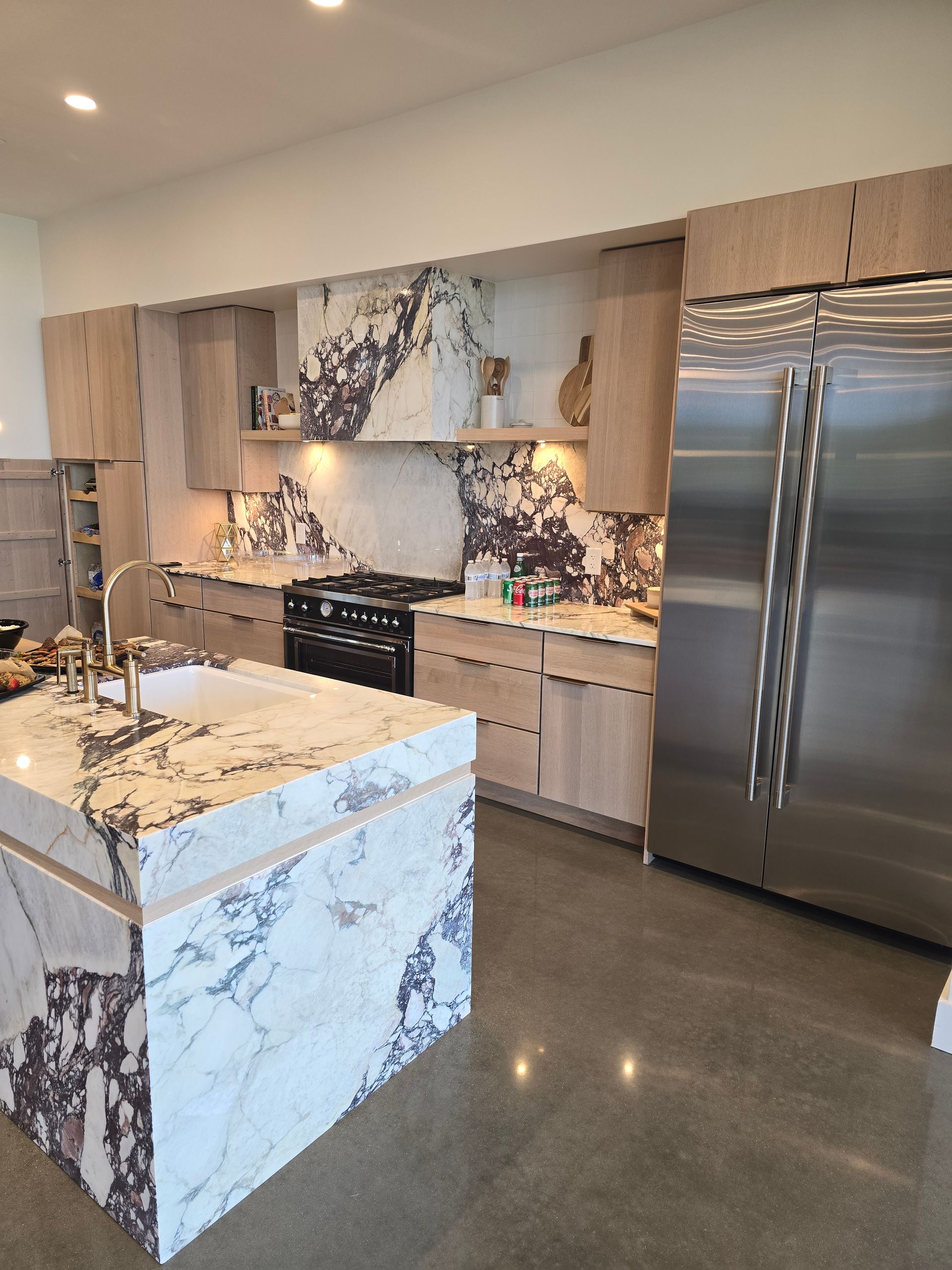 Modern kitchen with light wood cabinets, stainless steel refrigerator, and marble island.