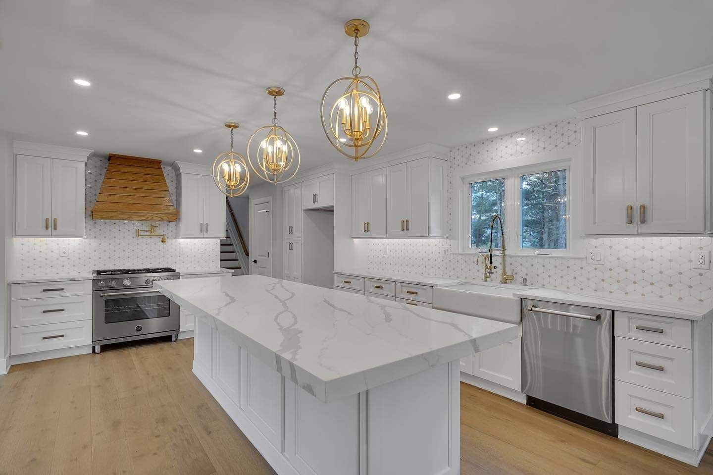 Modern white kitchen with marble countertops, stainless steel appliances, and gold light fixtures.