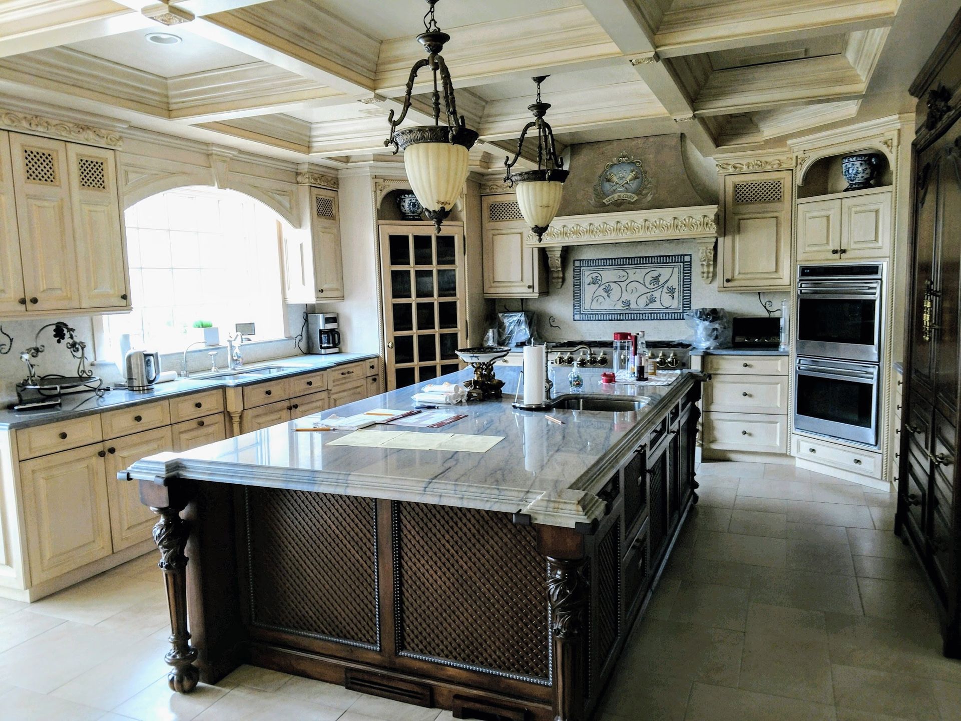 Luxurious cream-colored kitchen with large island, marble countertops, ornate cabinets, and hanging lights.
