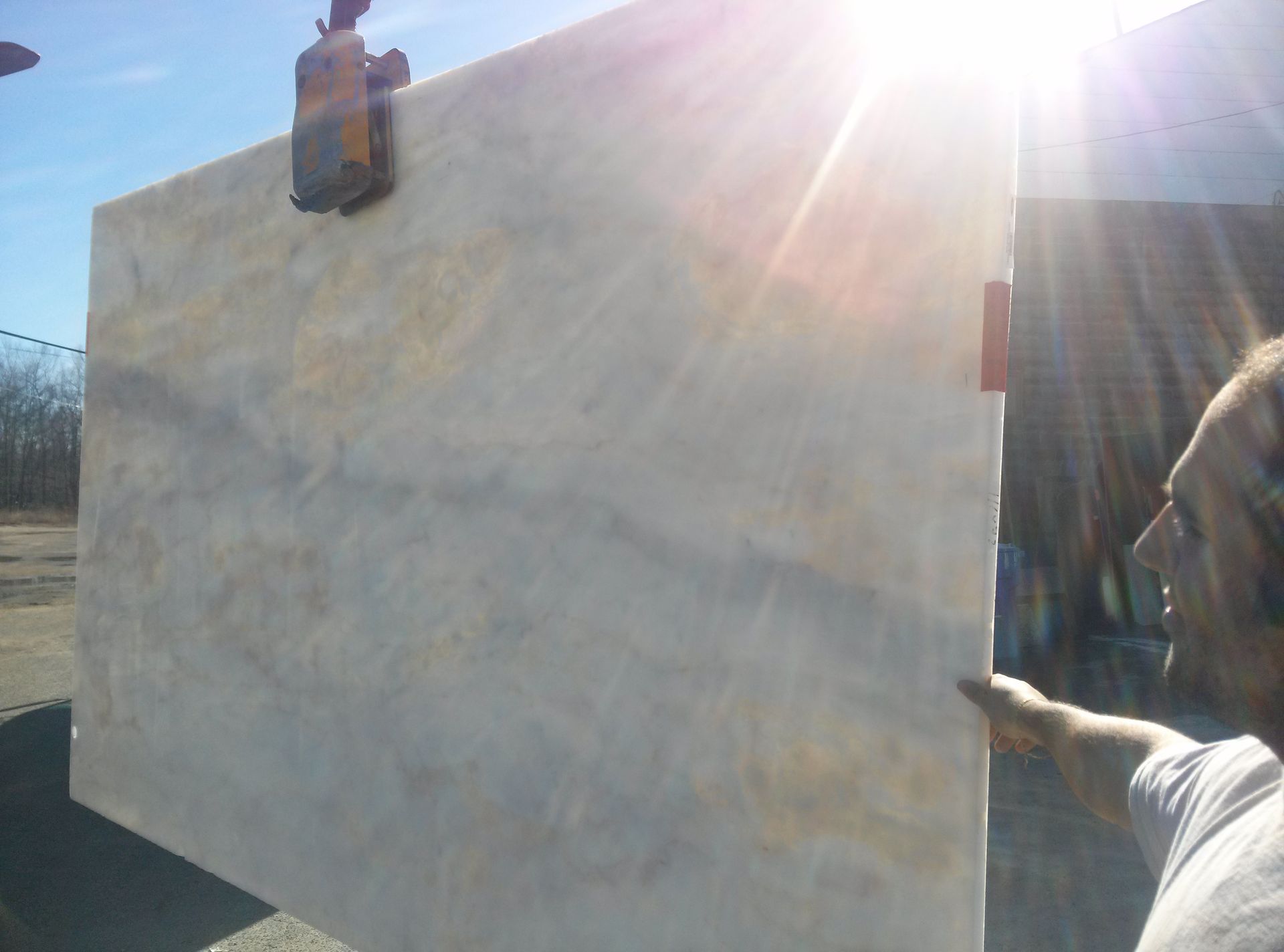 A man guides a large slab of stone lifted by machinery in bright sunlight.