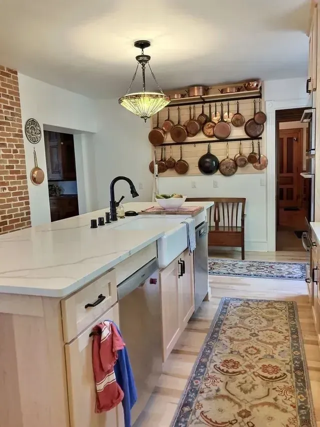 Kitchen with light cabinets, white countertop, brick wall, copper pots, and a long patterned rug.
