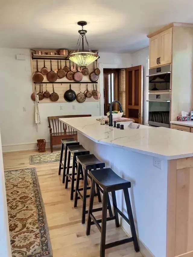 Kitchen with white countertop island, black stools, copper pots, and light wood cabinetry.
