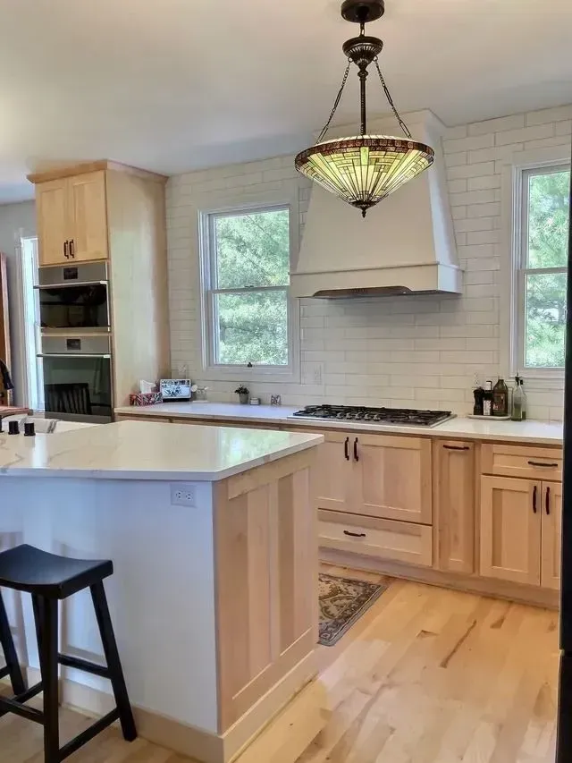 Light wood kitchen with island, white countertops, and subway tile backsplash. Decorative pendant light hangs above the cooktop.