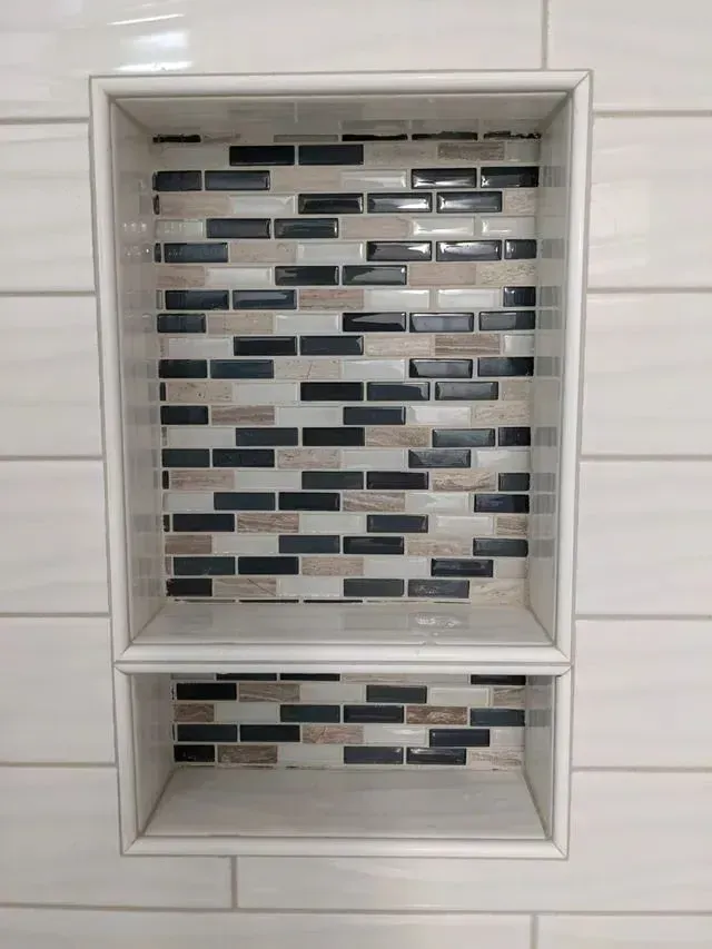 Recessed shower niche with shelf, tiled with black, gray, and beige mosaic tiles, set in white tile wall.