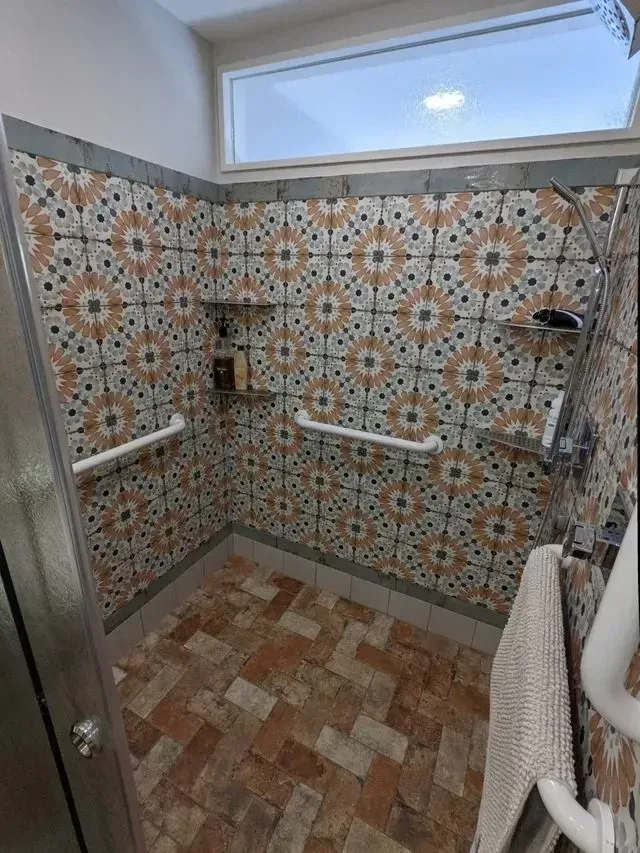 Shower with patterned orange, tan, and blue tiles, featuring grab bars, shelves, and a large window.