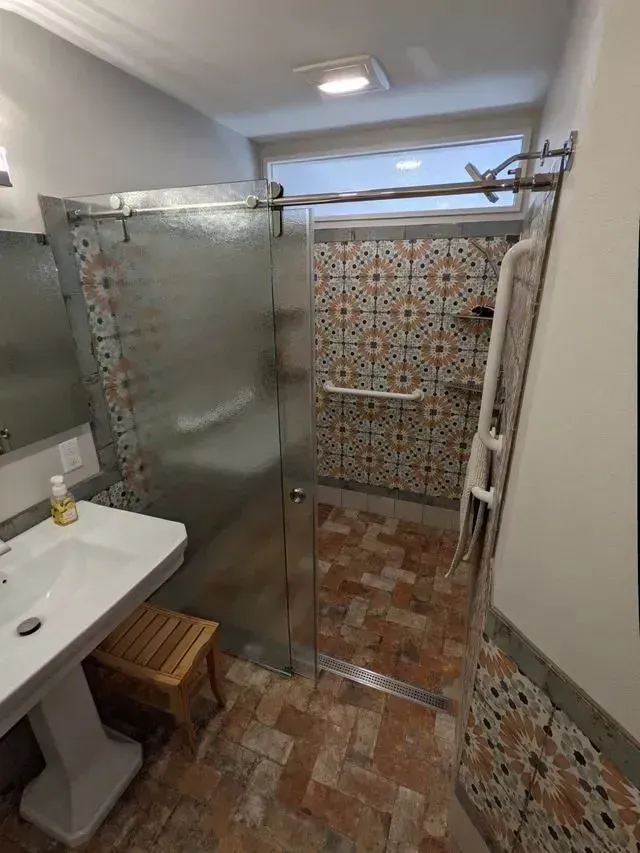 Bathroom with glass shower, patterned tile walls, and pedestal sink. Wooden stool.