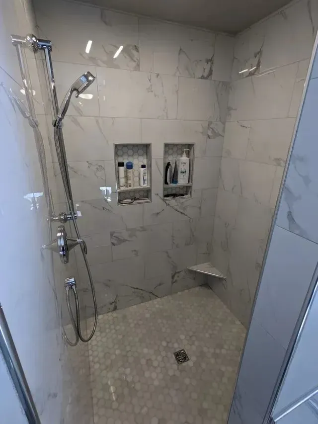 Modern white marble shower with two recessed shelves holding toiletries and a triangular seat.