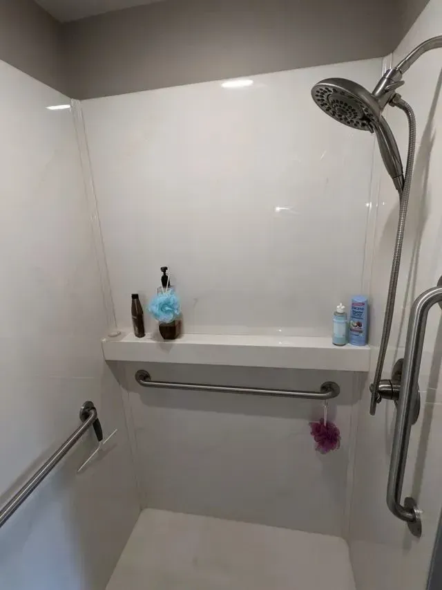 Shower stall with grab bars, built-in shelf with toiletries, and a showerhead. White walls.