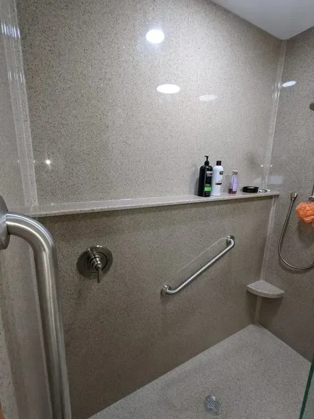 Accessible shower stall with grab bars, built-in shelf, beige speckled wall, and chrome fixtures.